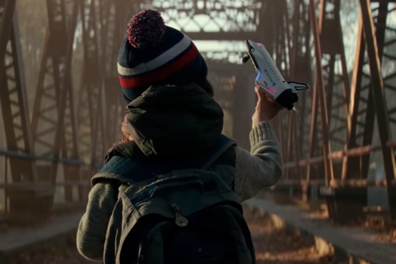 A shot of a little boy from behind. He stands in front of a bridge wearing a knit cap, vest, and backpack. His right hand is raised, and he holds a toy rocket.